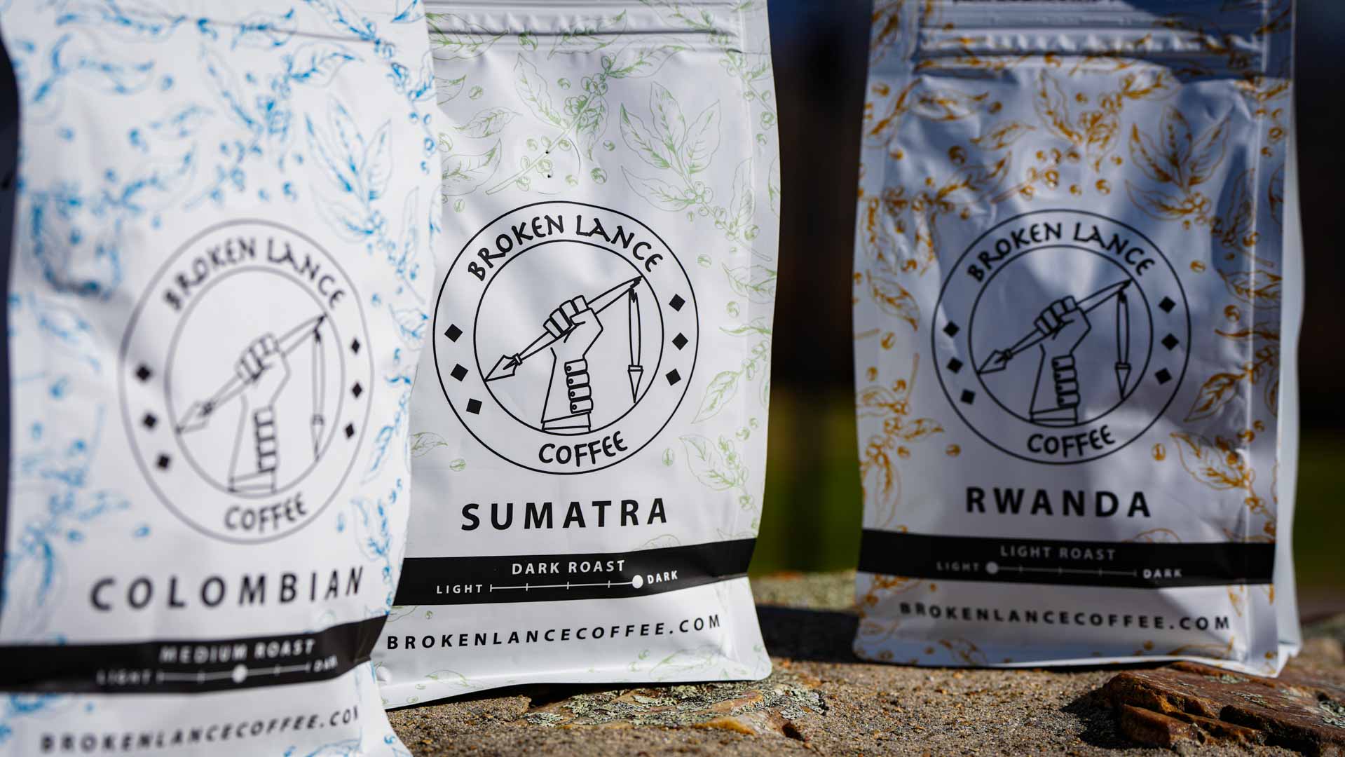 Three bags of Broken Lance Coffee are displayed outdoors in a scene perfect for photography & videography services. The Colombian, Sumatra, and Rwanda flavors each feature distinct color accents and roast levels shown at the bottom.