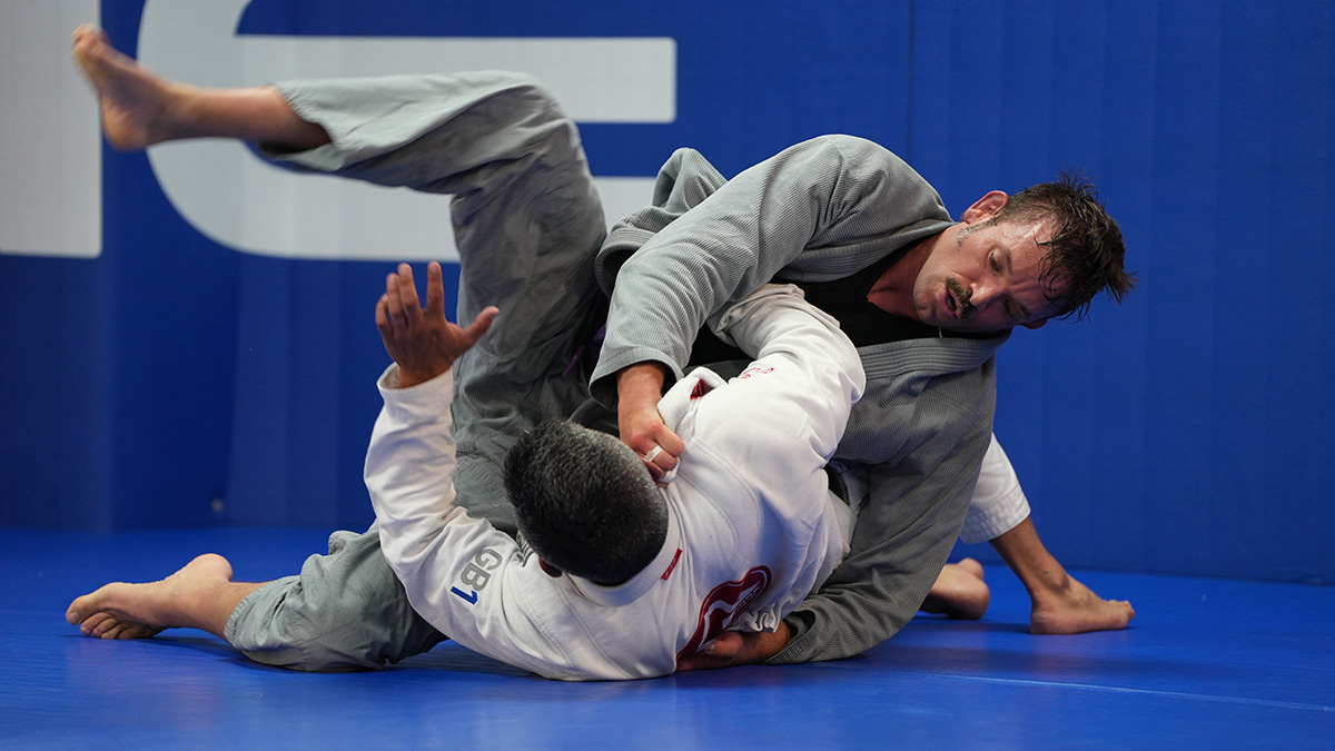 Two men in martial arts uniforms grapple on a blue mat during a Brazilian Jiu-Jitsu match; our photography & videography services capture every moment—one in a grey gi on top, the other in white defending from his back.