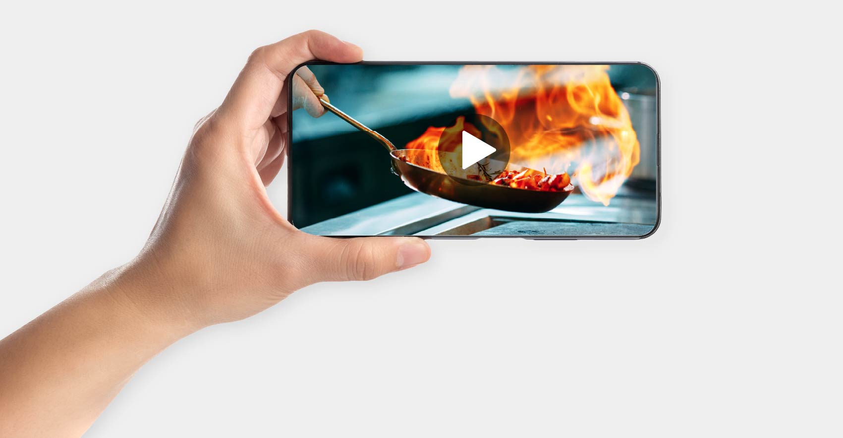 A hand holding a smartphone displaying a cooking video, showing food being flambéed in a pan with a large flame. The play button is visible on the phone screen.
