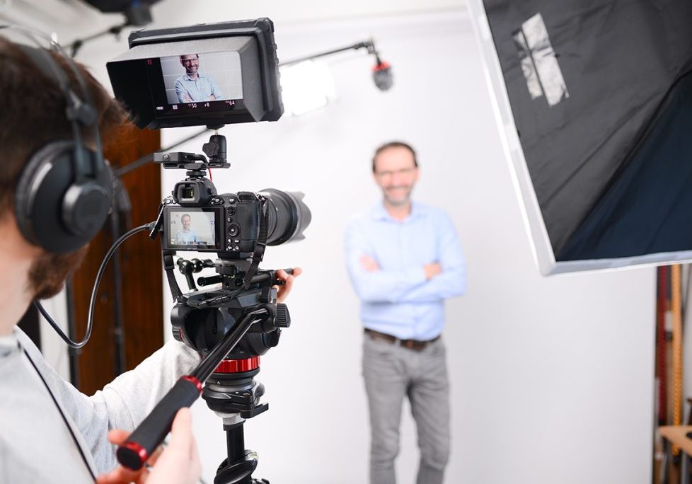 A person with headphones films a man standing with arms crossed in a studio setup, using a camera on a tripod. The man appears on the camera screen, and bright lighting equipment surrounds the scene.